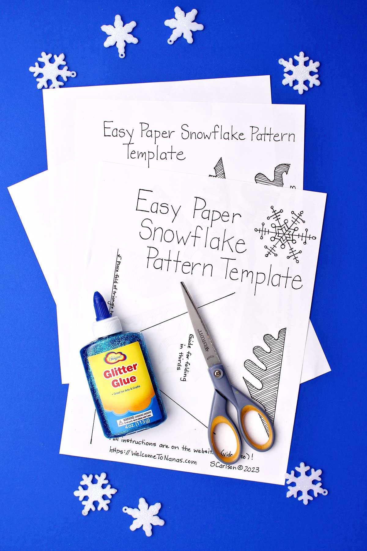 Easy Paper Snowflake Pattern Template, blue glitter glue and scissors on a blue background.