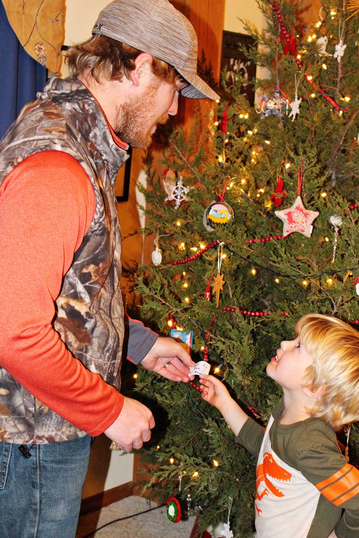 Man in camp vest and hat and young boy with blonde hair hold a clue in front of a decorated Christmas tree.