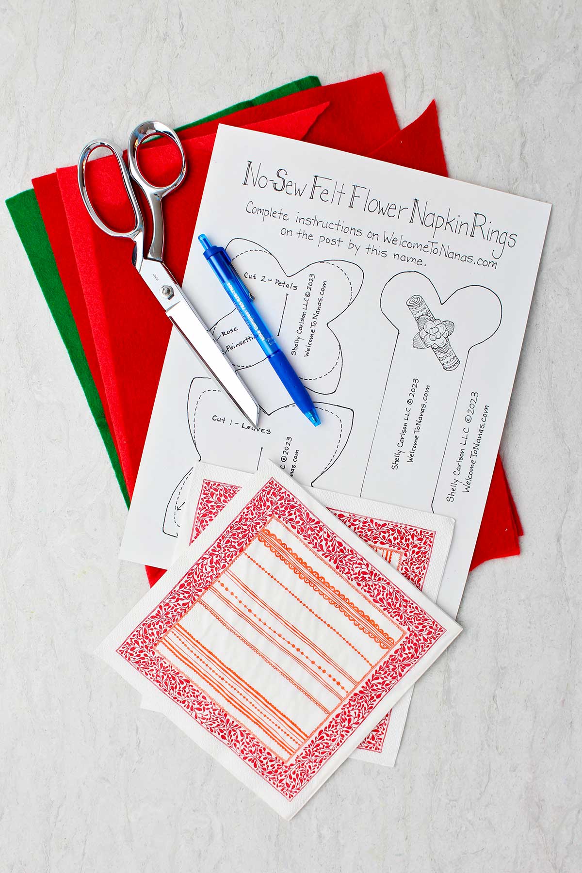 Supplies for a No-Sew Felt Christmas Napkin Ring. Pattern, scissors, pen, napkin and red and green felt.