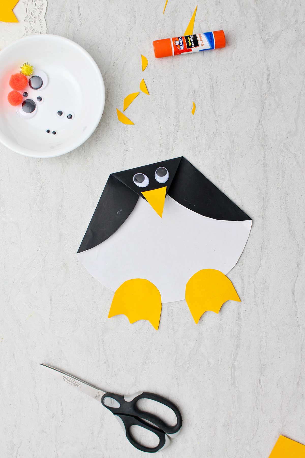 Completed Paper Penguin Craft with googly eyes and other supplies near by.