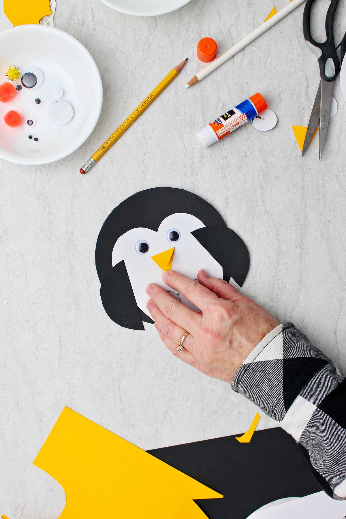 Hand gluing down beak in Paper Penguin Craft with holes for fingers with supplies near by.