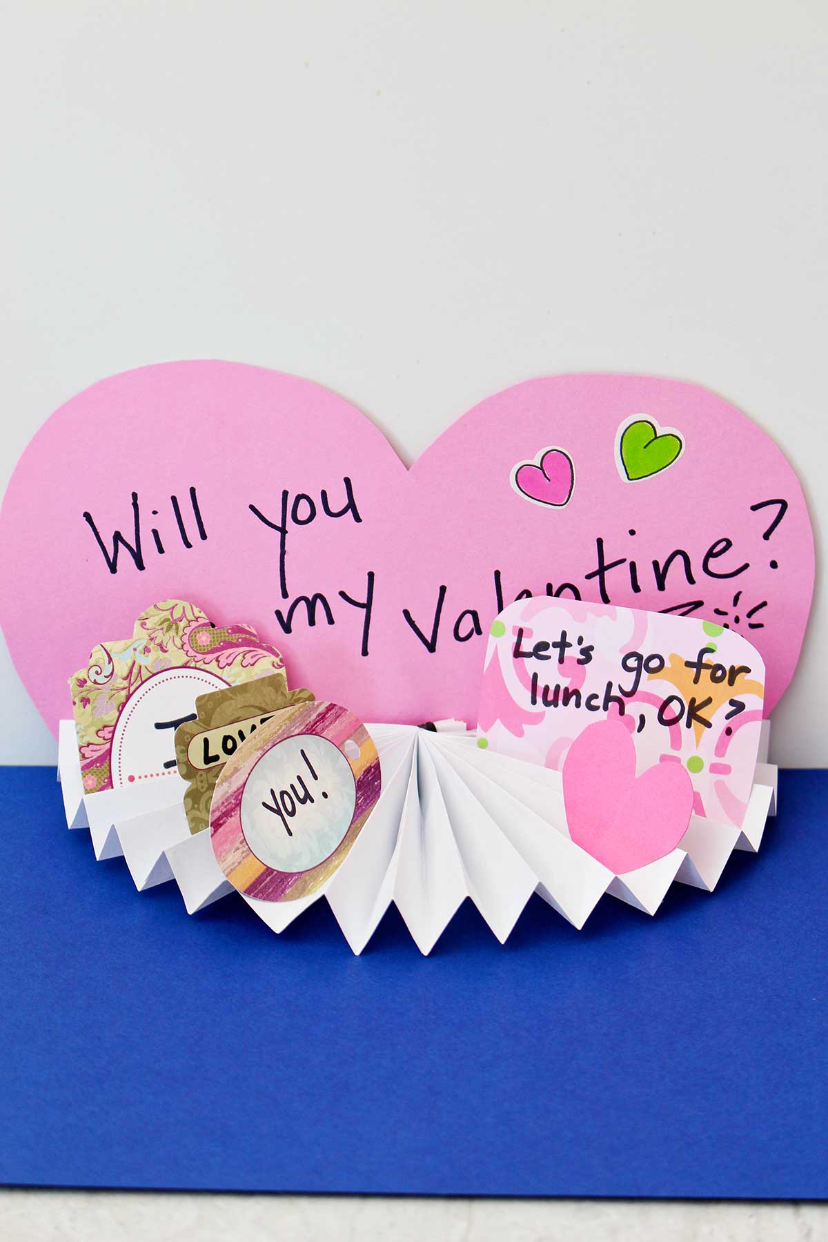 Completed Easy Paper Fan Craft in a Valentine's Day theme. Pink paper heart background says "Will You Be My Valentine" with other cute notes in the fan.