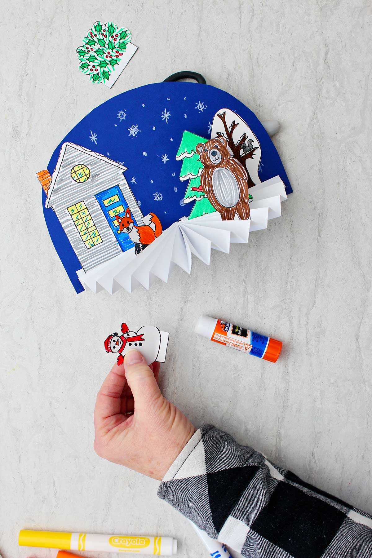 Hand holding snowman cutout about to be glued to the winter themed Easy Paper Fan Craft.