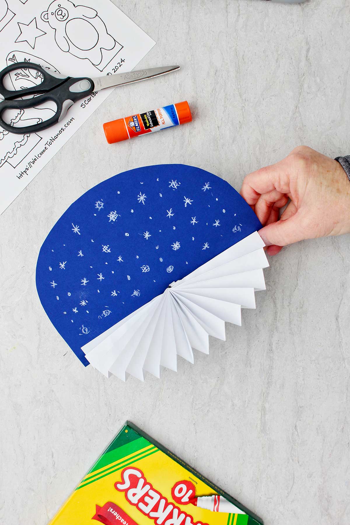 Hand holding blue snowy background with white fan glued to the bottom for winter theme.