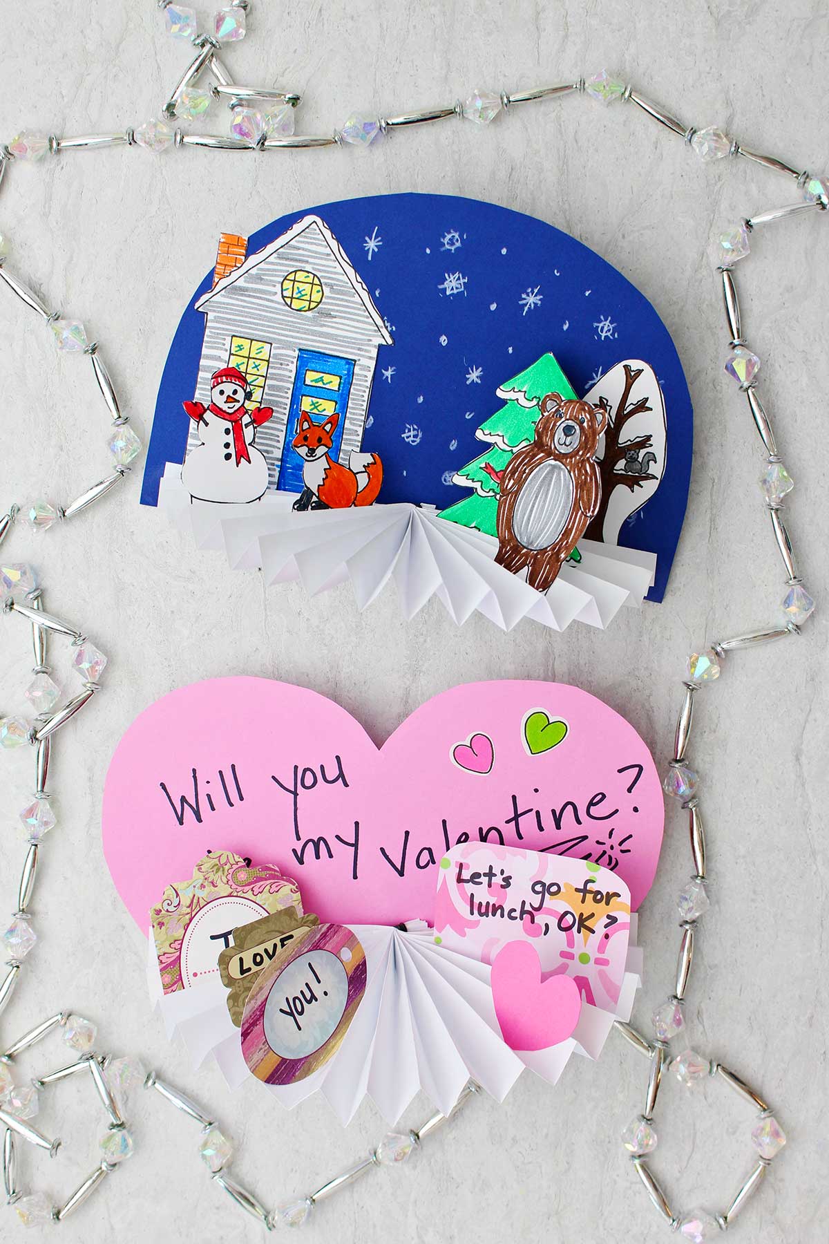 Two images of completed Paper Fan Crafts with beads around it. One with a winter theme and one with a Valentine's Day theme.