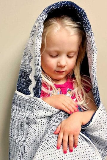 Young girl with blonde hair snuggled up in a hooded blue and white towel looking down.