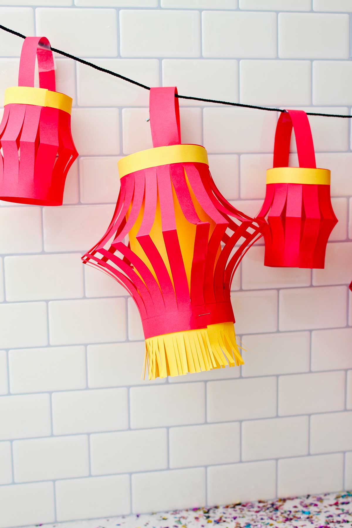 Three completed paper lanterns hanging against a subway tile backdrop.