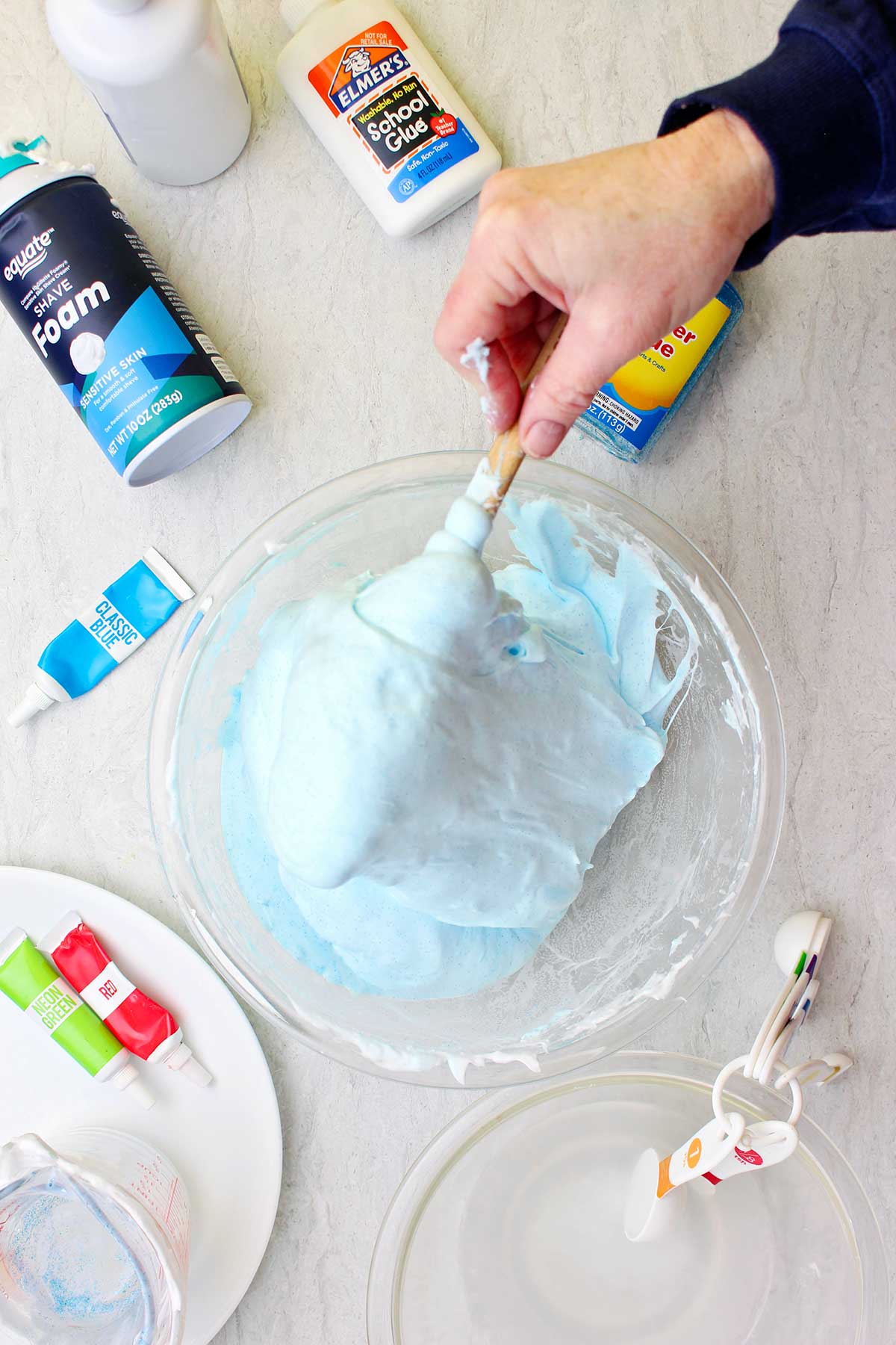 Hand stirring a batch of light blue fluffy slime in a glass bowl with supplies scattered near by.