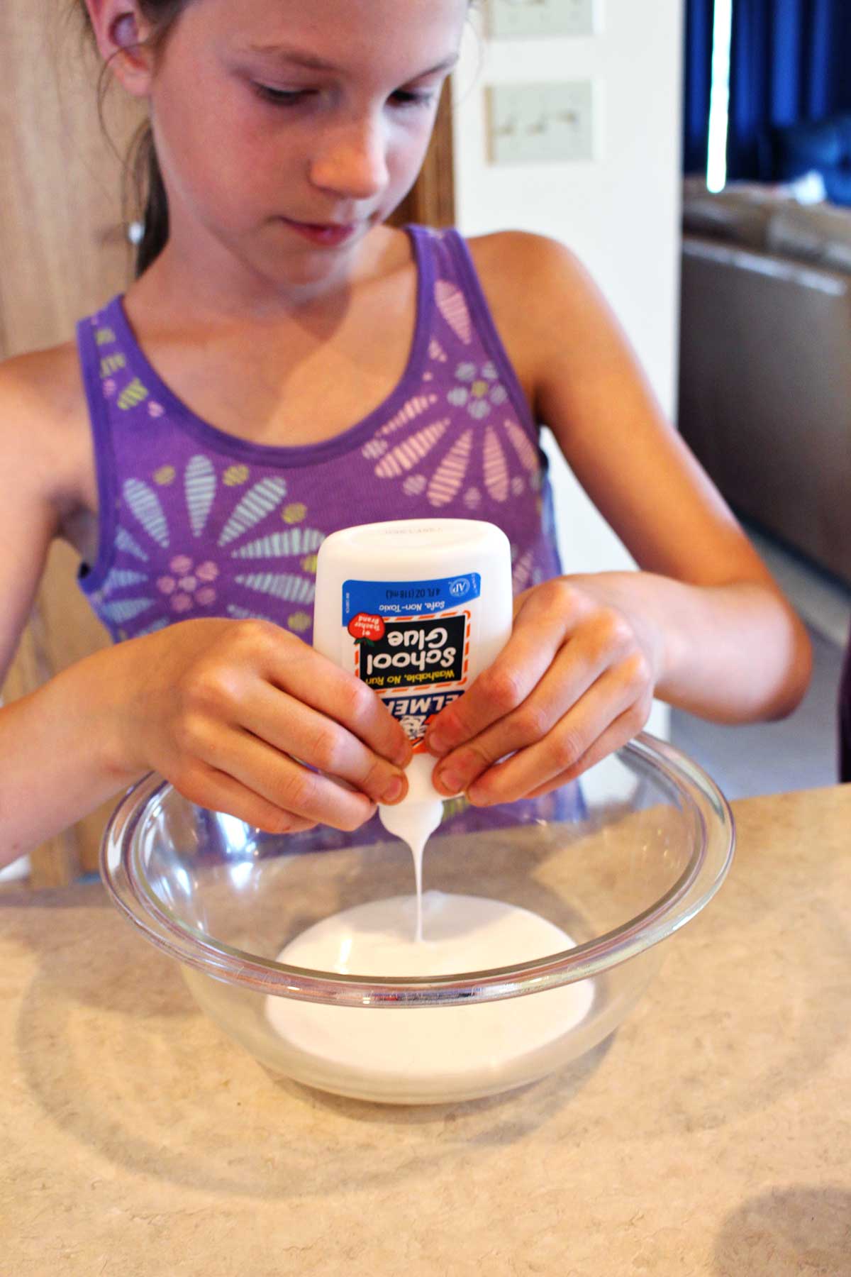 Girl in purple tank top adds Elmer's Glue to a glass bowl.