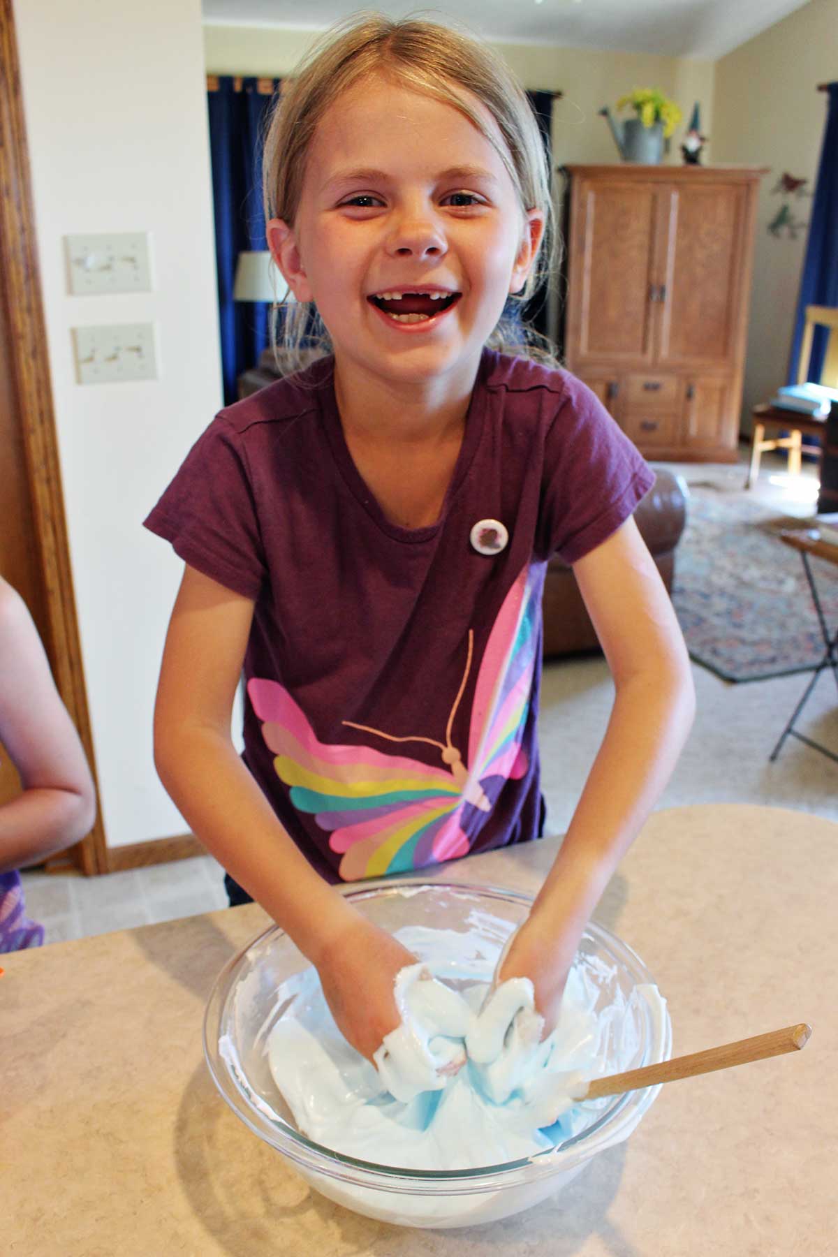 Young girl in maroon butterfly shirt smiling at the camera playing with batch of fluffy slime in a glass bowl.