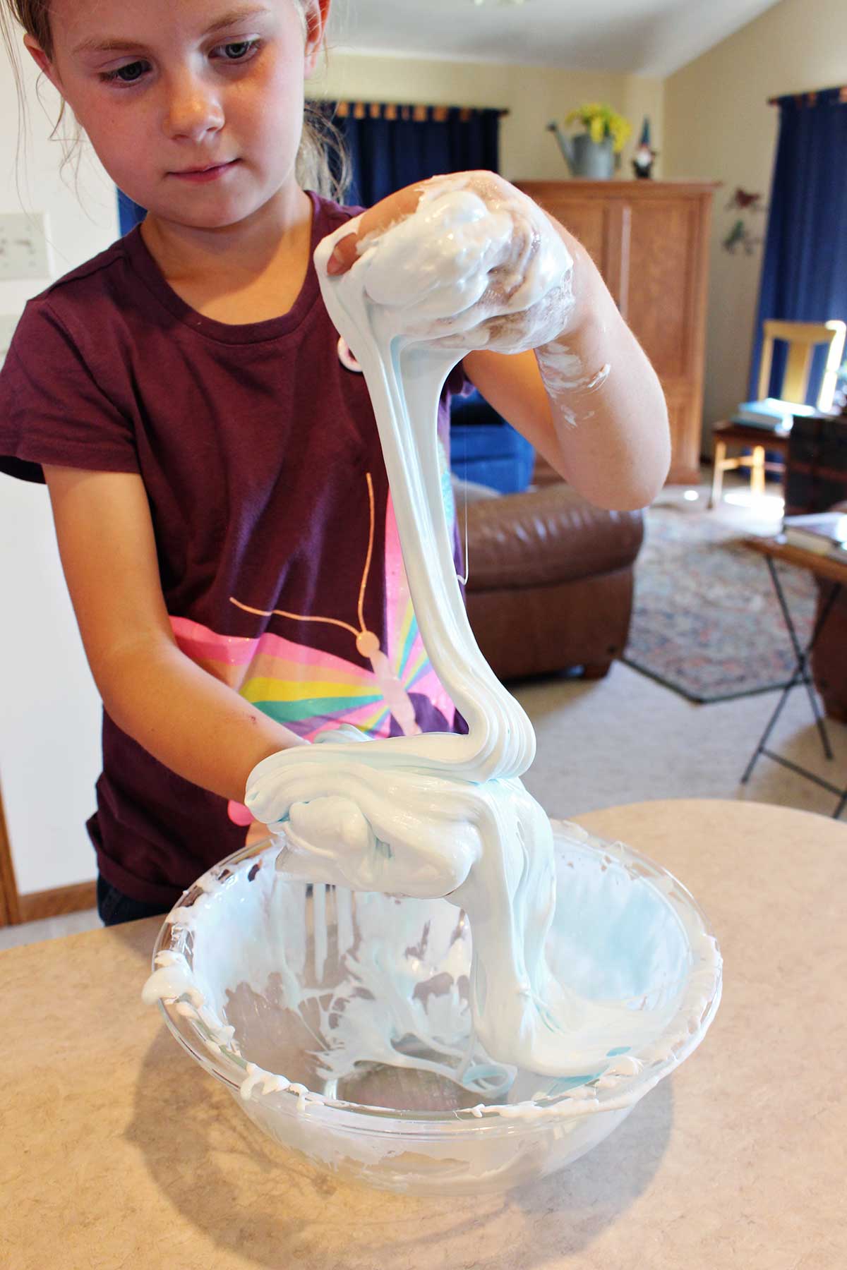Young girl in maroon butterfly shirt playing with batch of fluffy slime in a glass bowl.