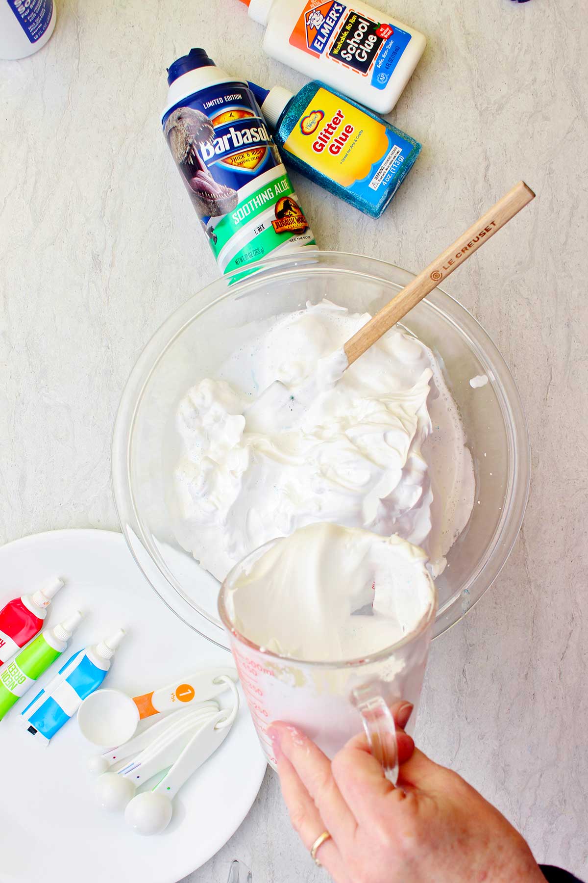 Hand pouring an ingredient into a bowl of shaving cream in a glass bowl with supplies for fluffy slime scattered near by.