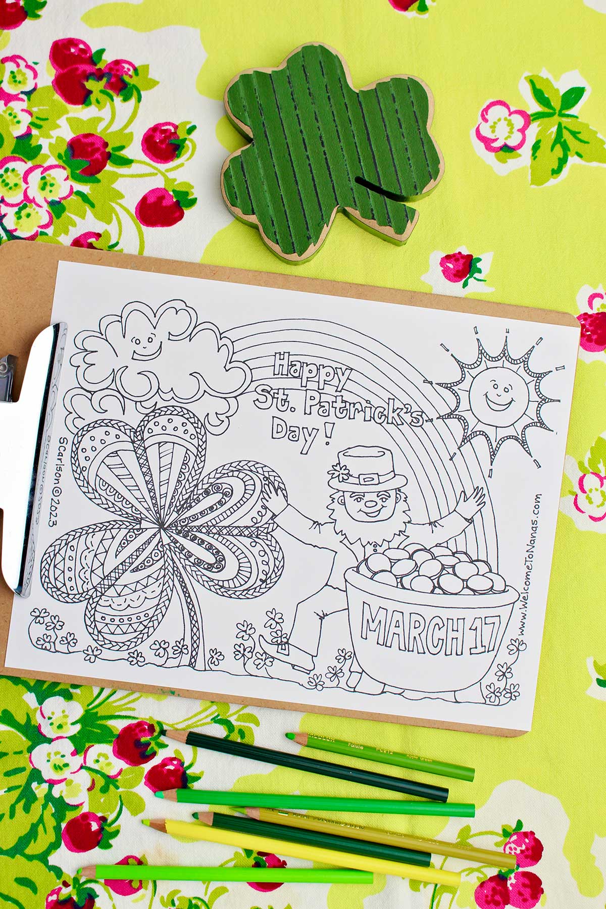 Uncolored St. Patrick's Day coloring page on a clip board resting on a green table cloth with strawberries on it with green colored pencils and a wooden shamrock near by.