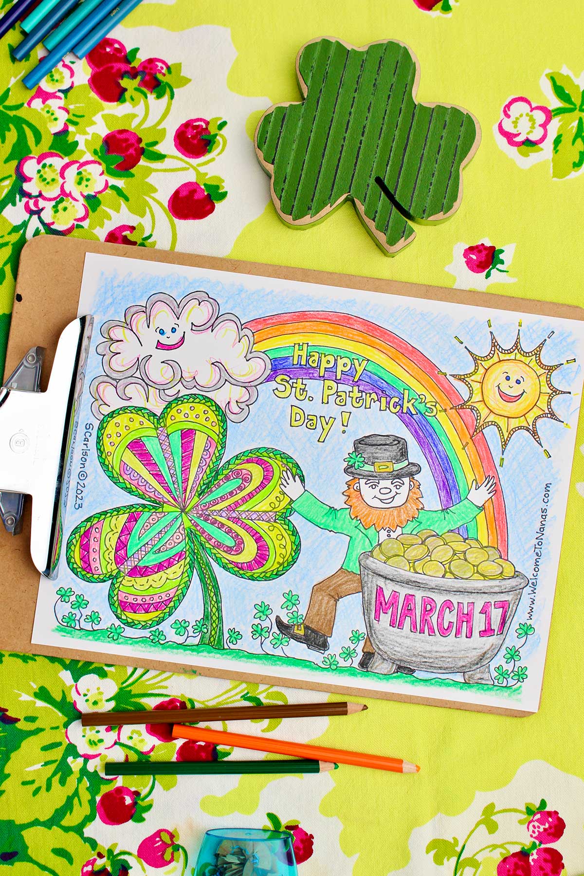 Completed St. Patrick's Day coloring page on clipboard sitting on a green table cloth with strawberries on it with a wooden shamrock and colored pencils near by.