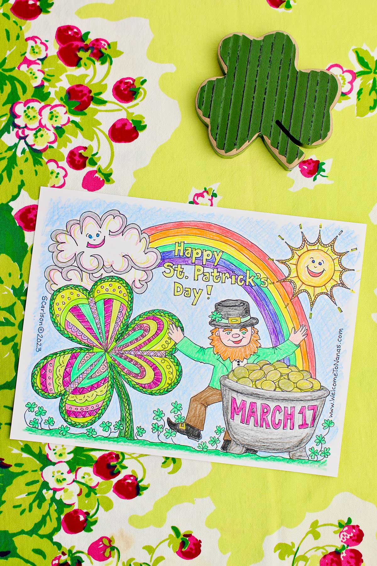 Completed St. Patrick's Day coloring page sitting on a green table cloth with strawberries on it with wooden shamrock near by.
