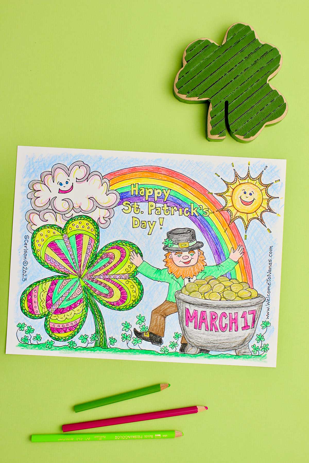 Vertical image of completed St. Patrick's Day coloring page against a lime green backdrop with three colored pencils and a wooden shamrock near by.