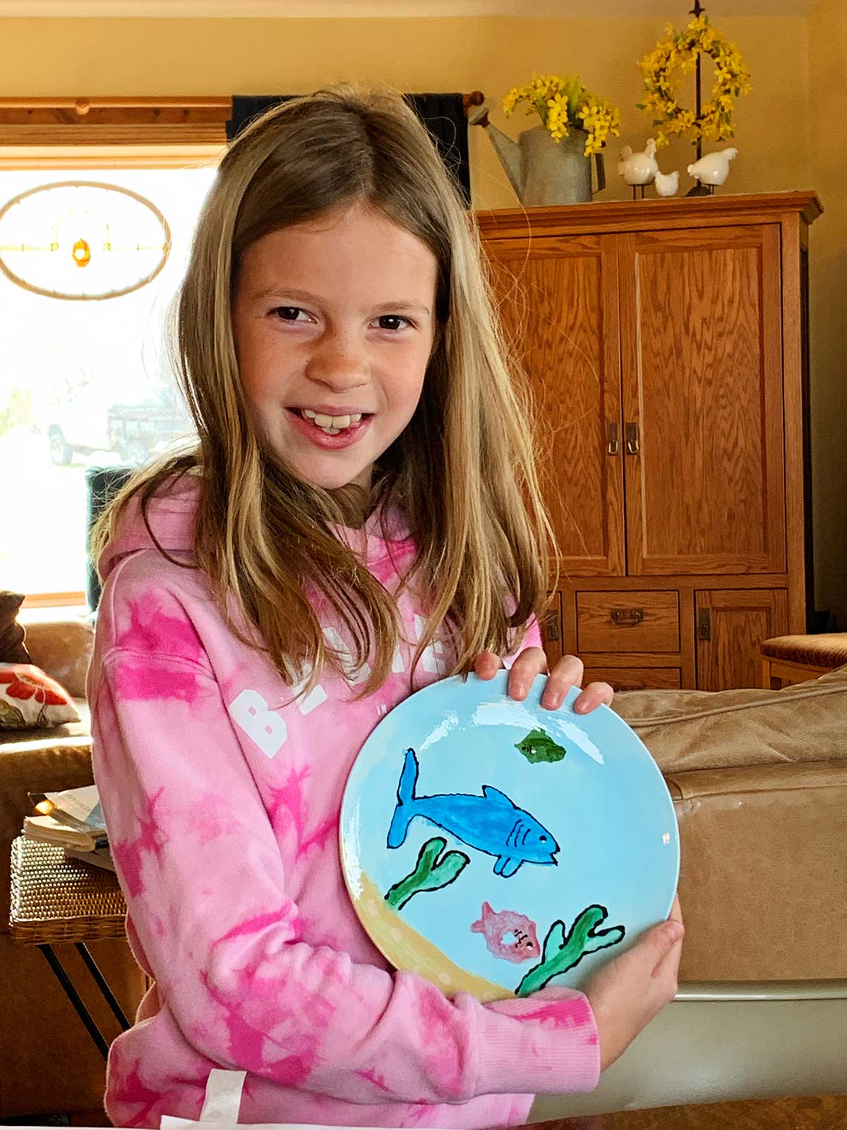 Young girl with light brown hair and brown eyes wearing a pink sweatshirt shows off her completed pottery project of a plate with a shark and ocean scene painted on it.