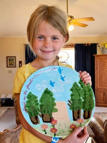 Young girl with blonde hair and blue eyes and a yellow shirt holds her completed pottery project of a plate showing a nature scene.