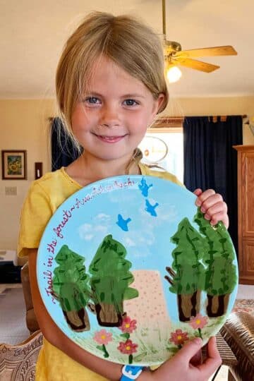 Young girl with blonde hair and blue eyes and a yellow shirt holds her completed pottery project of a plate showing a nature scene.