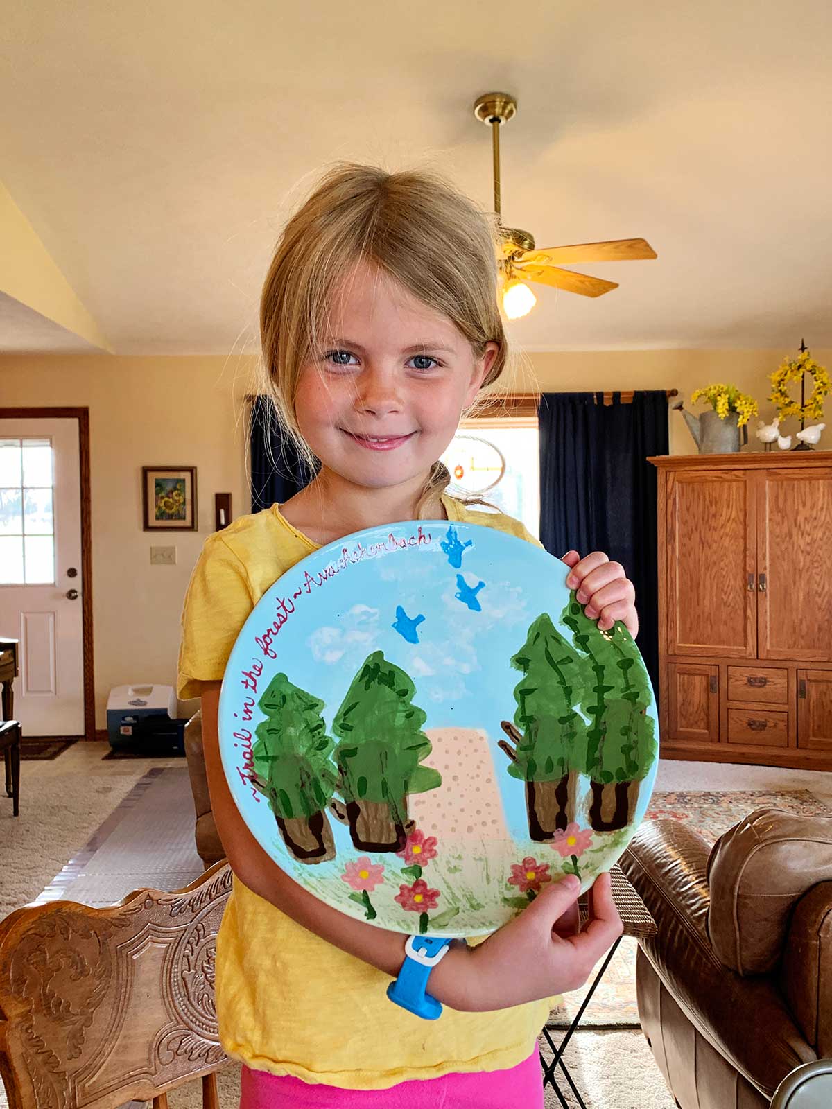 Young girl with blonde hair and blue eyes and a yellow shirt holds her completed pottery project of a plate showing a nature scene.