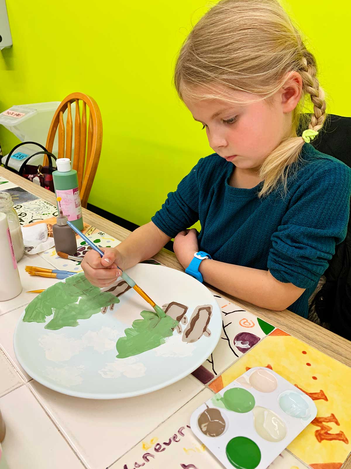 Young girl with blonde hair and braids paints her plate at ceramic painting studio.