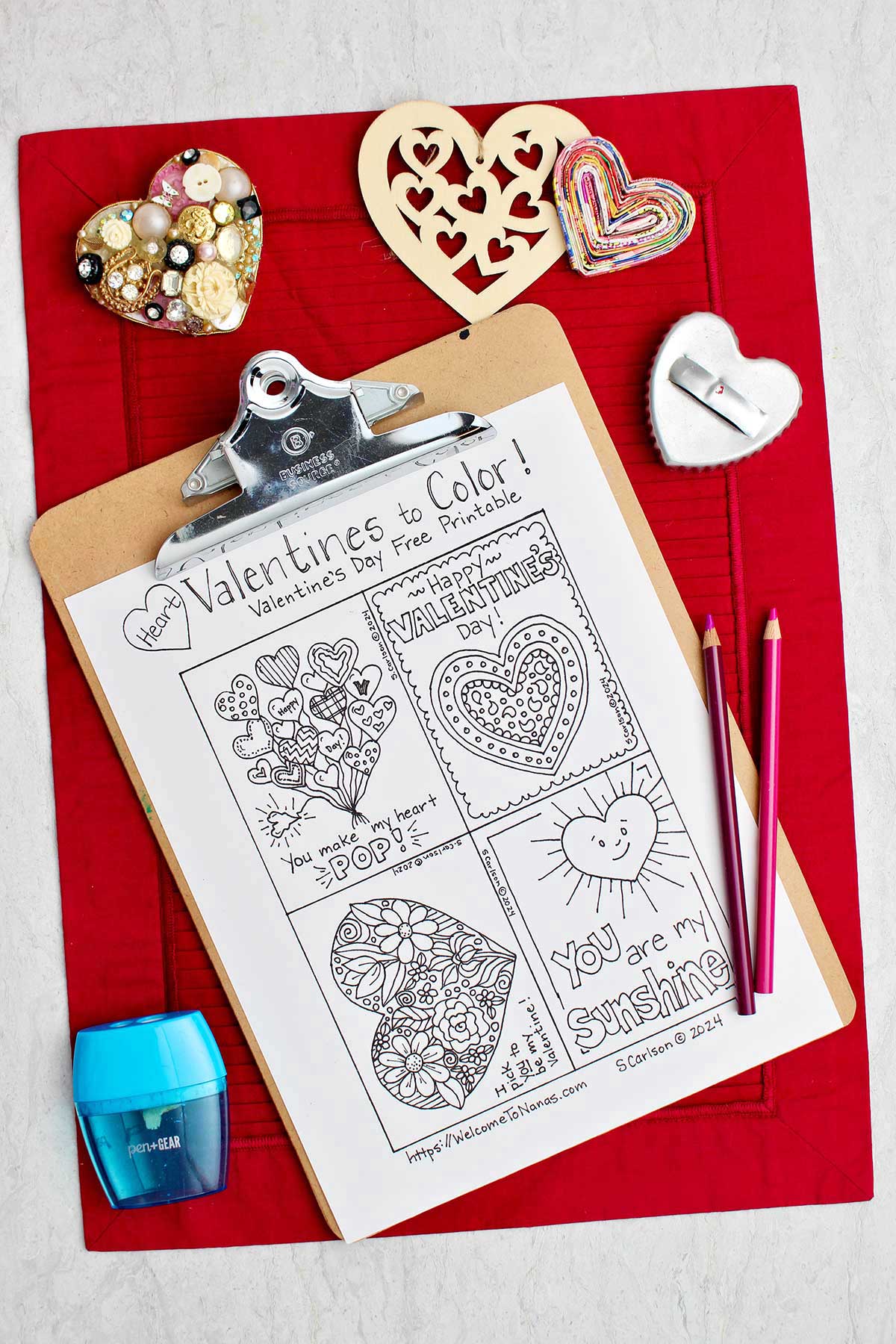Uncolored Valentine's Day printable on a clip board resting on a red placemat with heart shaped objects and colored pencils resting near by.