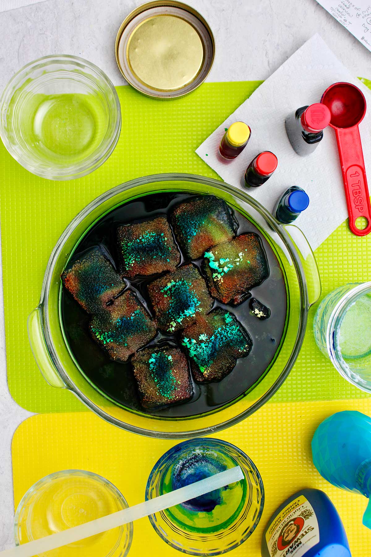Beginning process of teal and blue growing crystals on charcoal blocks in glass tray sitting on green plastic cutting board with supplies near by.