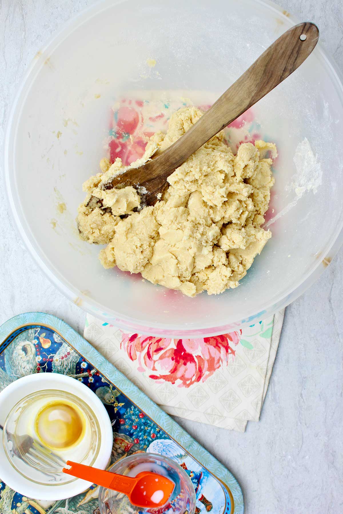 Bowl of sugar cookie dough with wooden spoon resting in it with tray of other ingredients near by.