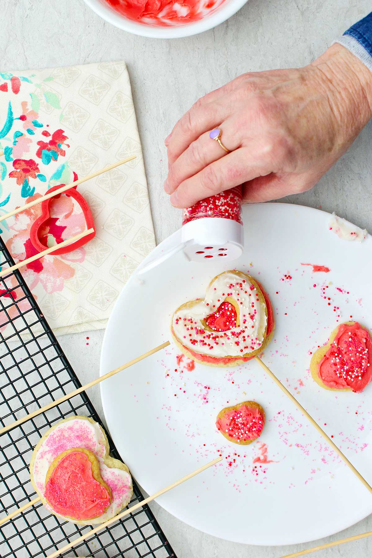 Hand sprinkling red and white sprinkled on iced cookie on a stick.