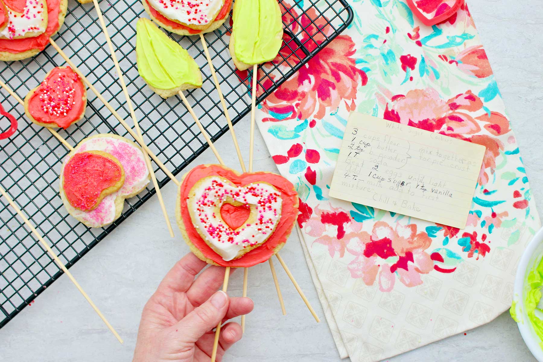Hand holding iced sugar cookie on a stick with other completed cookies in the background.