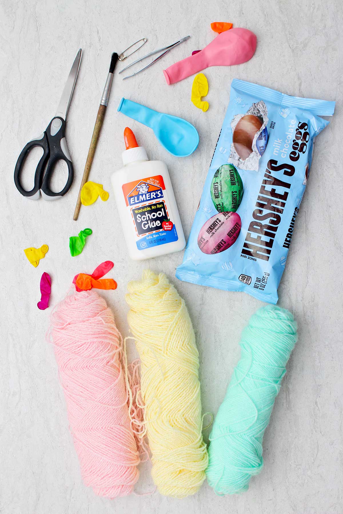 Supplies for String Eggs for Easter. Three colors of yarn, small balloons, white glue, scissors, paintbrush, tweezers and candy eggs.