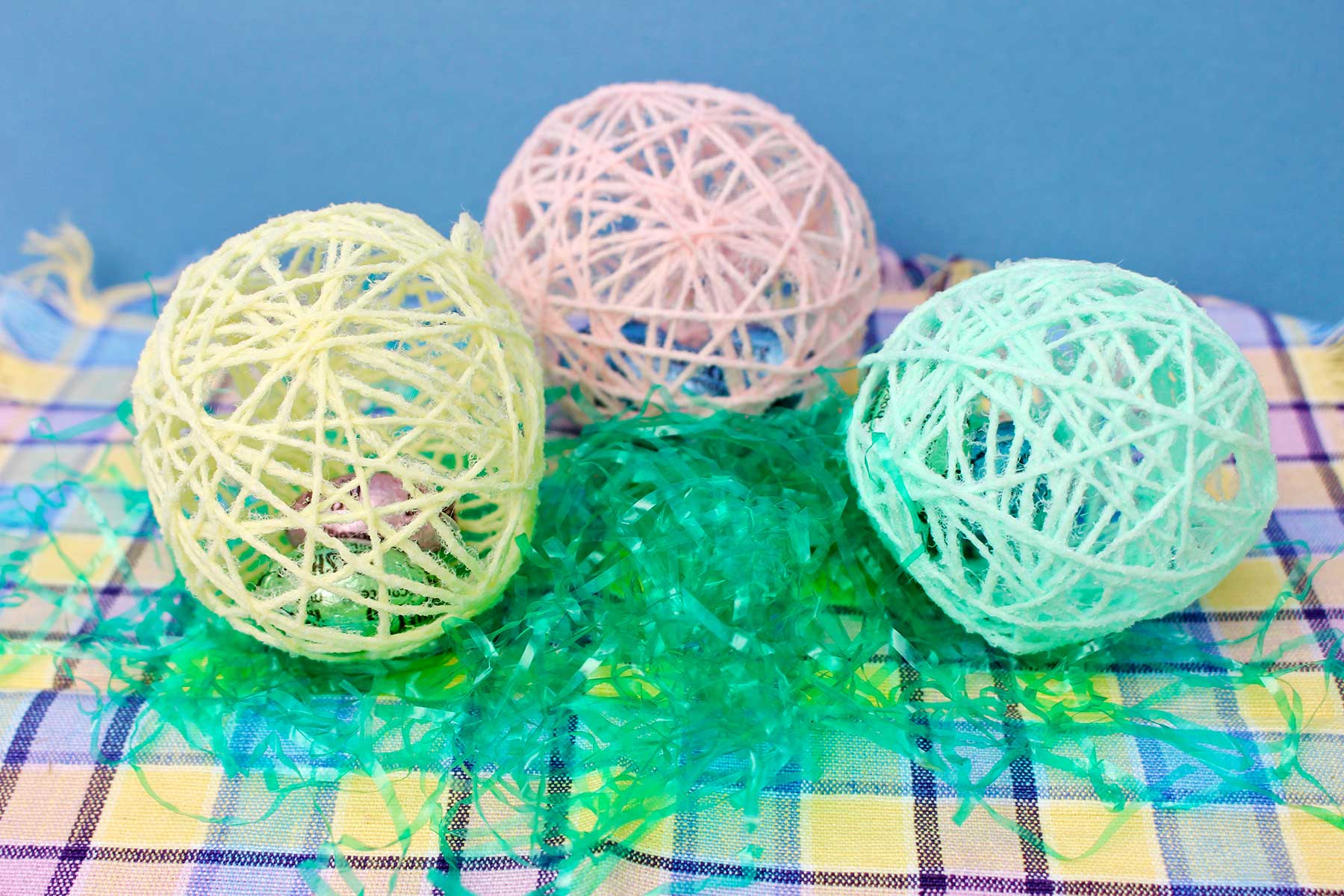 Yellow, pink and green string eggs sitting on top of Easter basket grass on a colorful tablecloth.