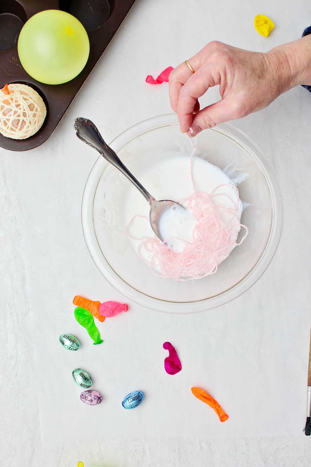 Hand dipping pink yarn in a bowl of white glue with other supplies near by.