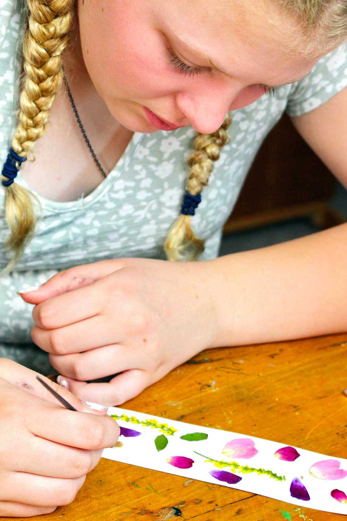 Young girl with blonde braids arranges flower petals on a strip of paper.