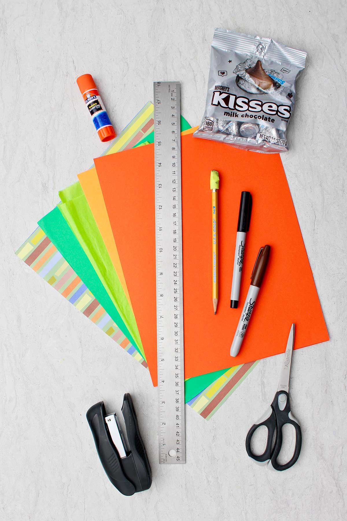 Supplies to make carrot Easter baskets. Colorful paper, glue stick, candy, scissors, pencil, markers, stapler and ruler.