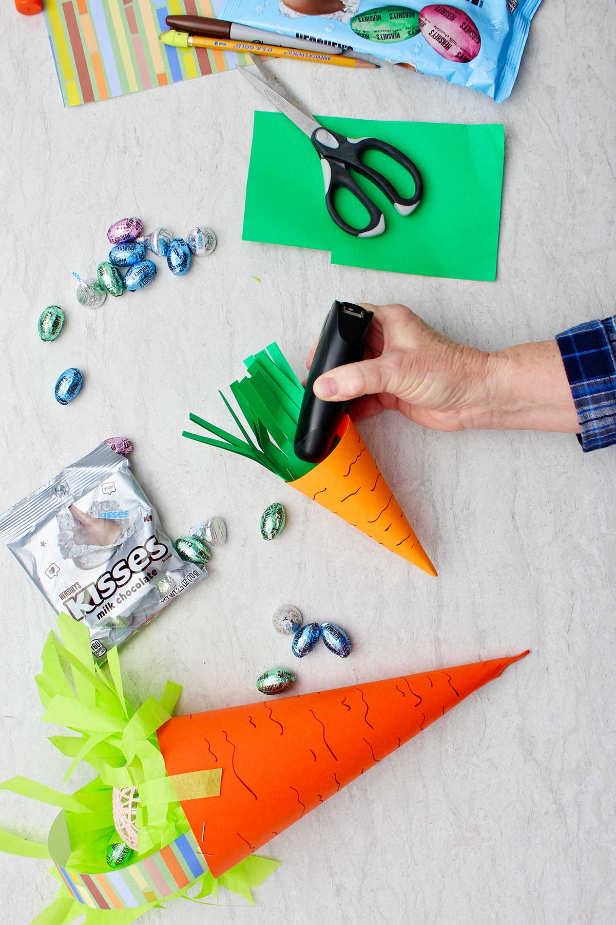 Hand stapling green paper grass to inside of smaller carrot Easter basket with other supplies near by.