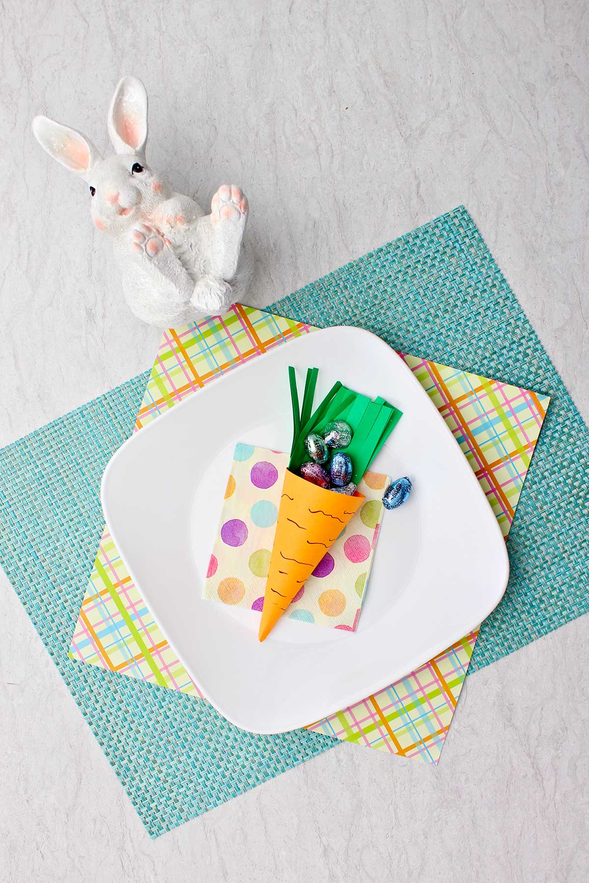 Smaller carrot Easter basket sitting on a white plate, colorful plaid piece of paper and robins egg blue placemat filled with paper grass and easter candy with white bunny statue above it.