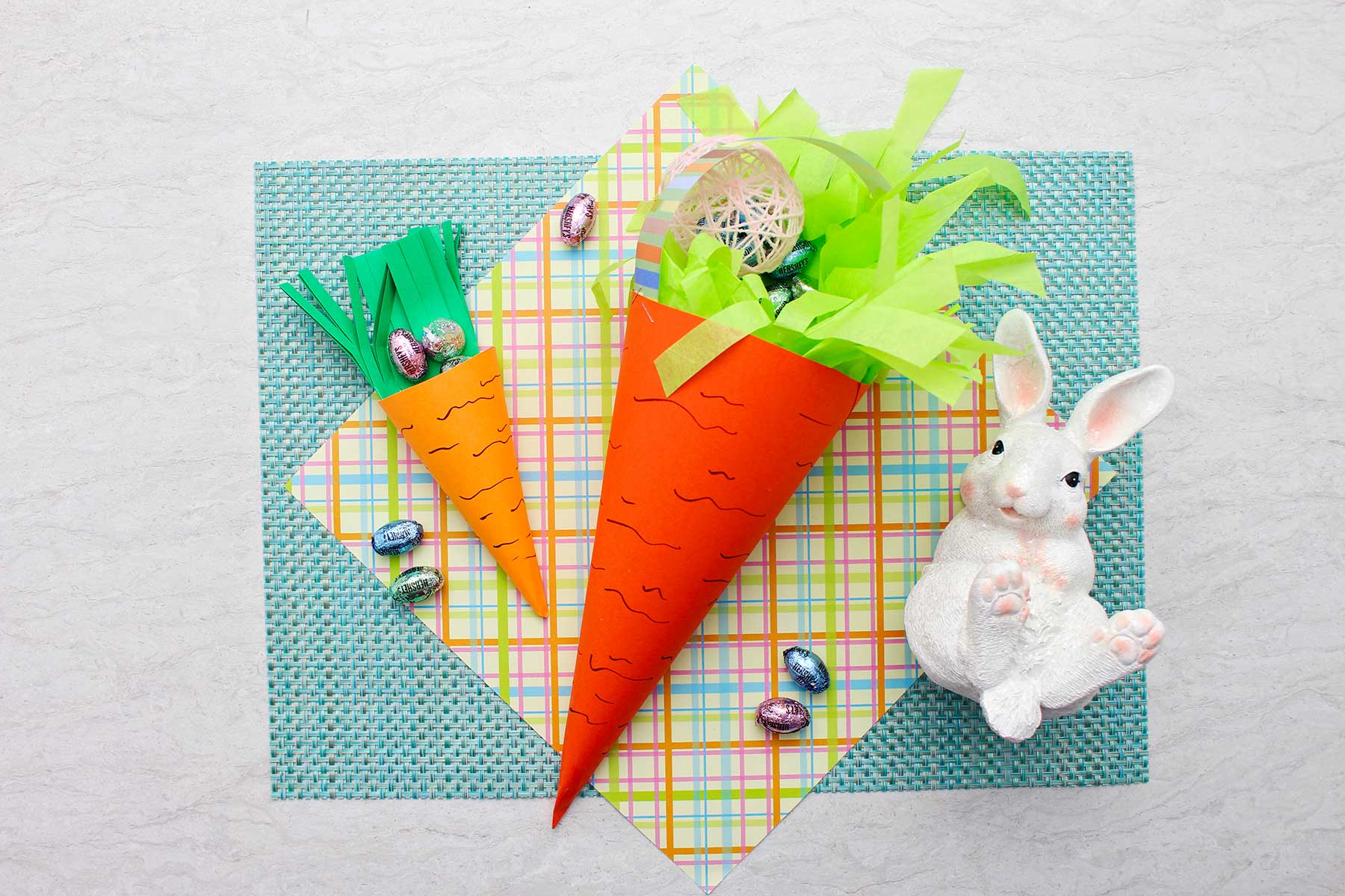 Completed carrot Easter baskets, one large and one smaller filled with paper grass and easter egg candy on a plaid piece of paper and robins egg blue placemat.