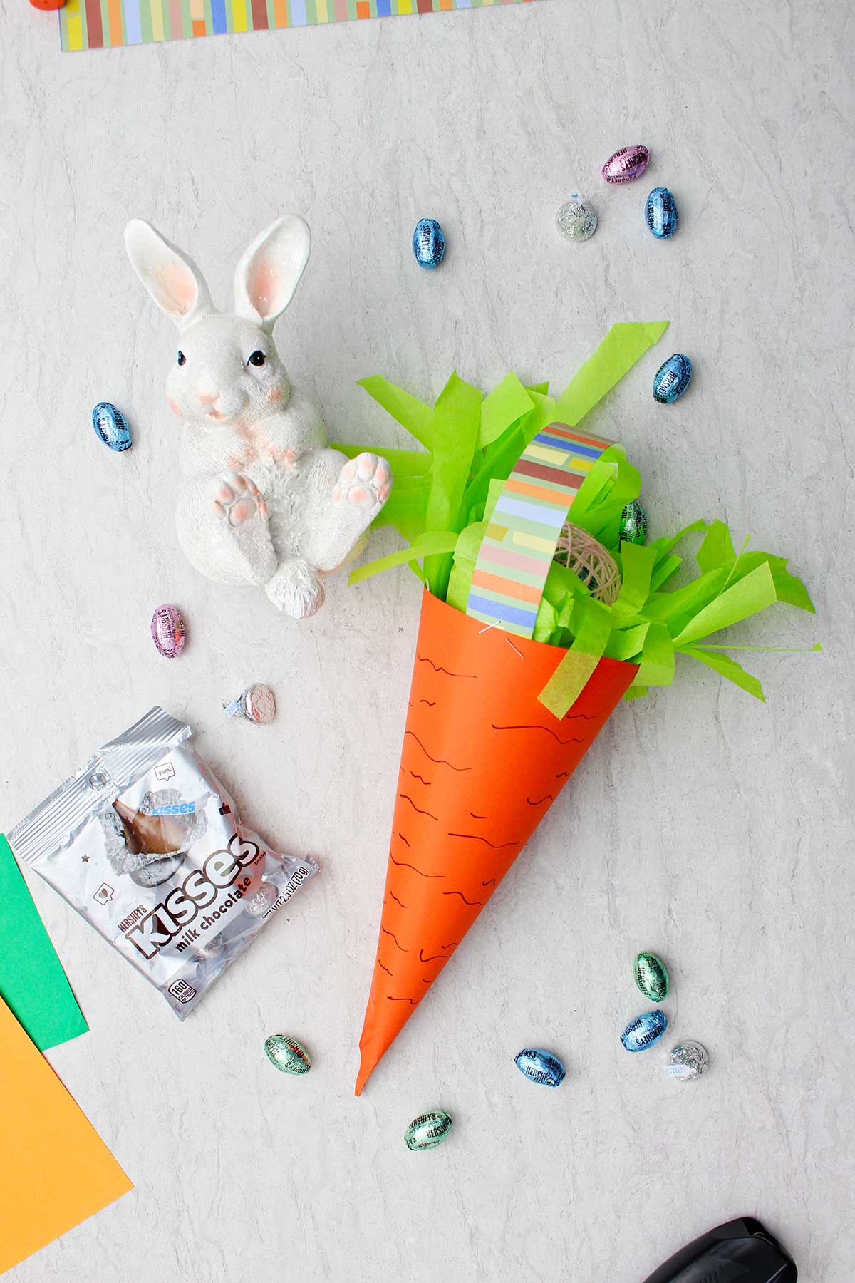 Completed large carrot Easter basket with paper grass and Easter candy inside with white bunny statue, candies and paper near by.
