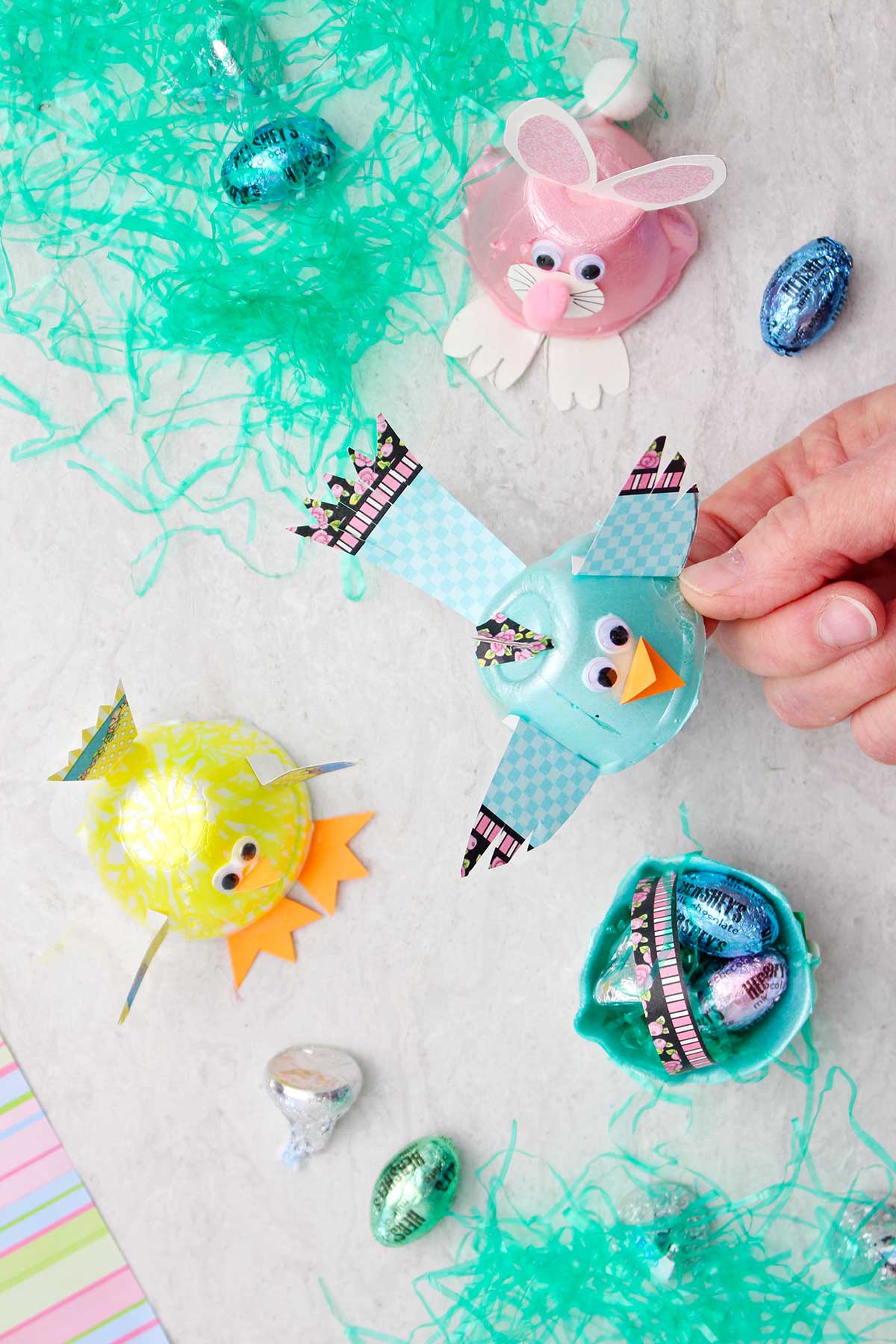 Hand holding a blue bird egg carton animal with other completed animals, Easter basket grass and candy in the background.
