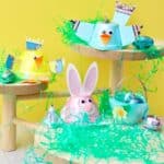 Three egg carton animals sitting on wooden stands with Easter basket grass and candy around them against a yellow backdrop.