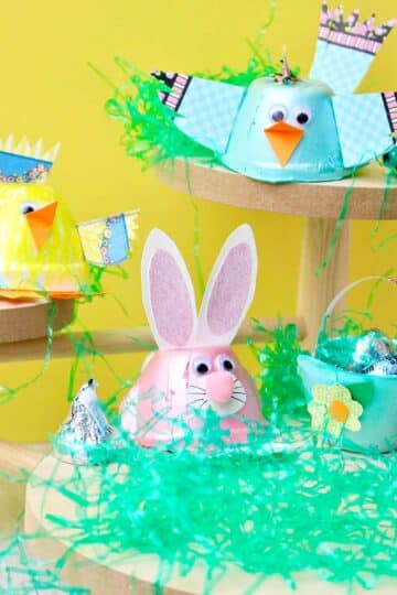 Three egg carton animals sitting on wooden stands with Easter basket grass and candy around them against a yellow backdrop.