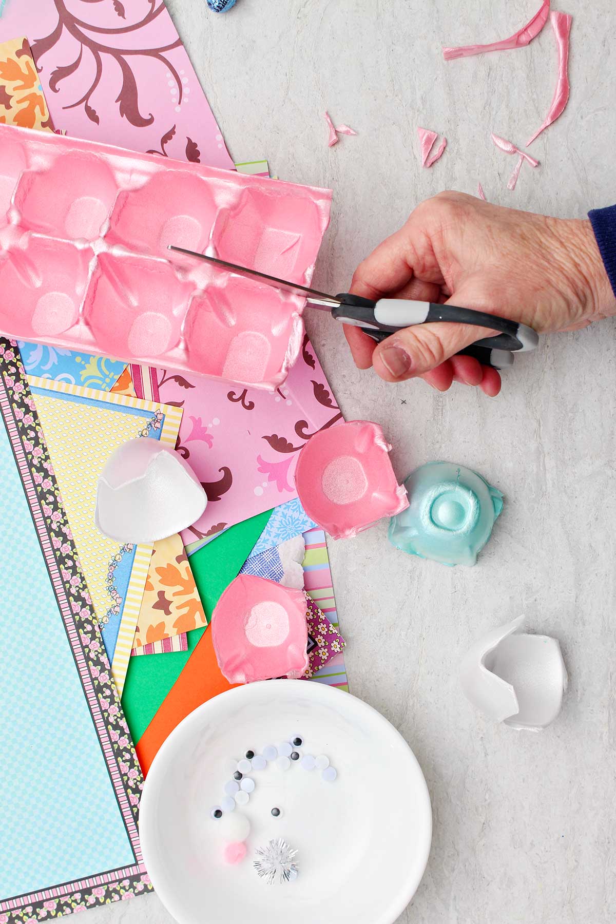 Hand cutting up a pink styrofoam egg carton with scraps of colorful paper in the background.