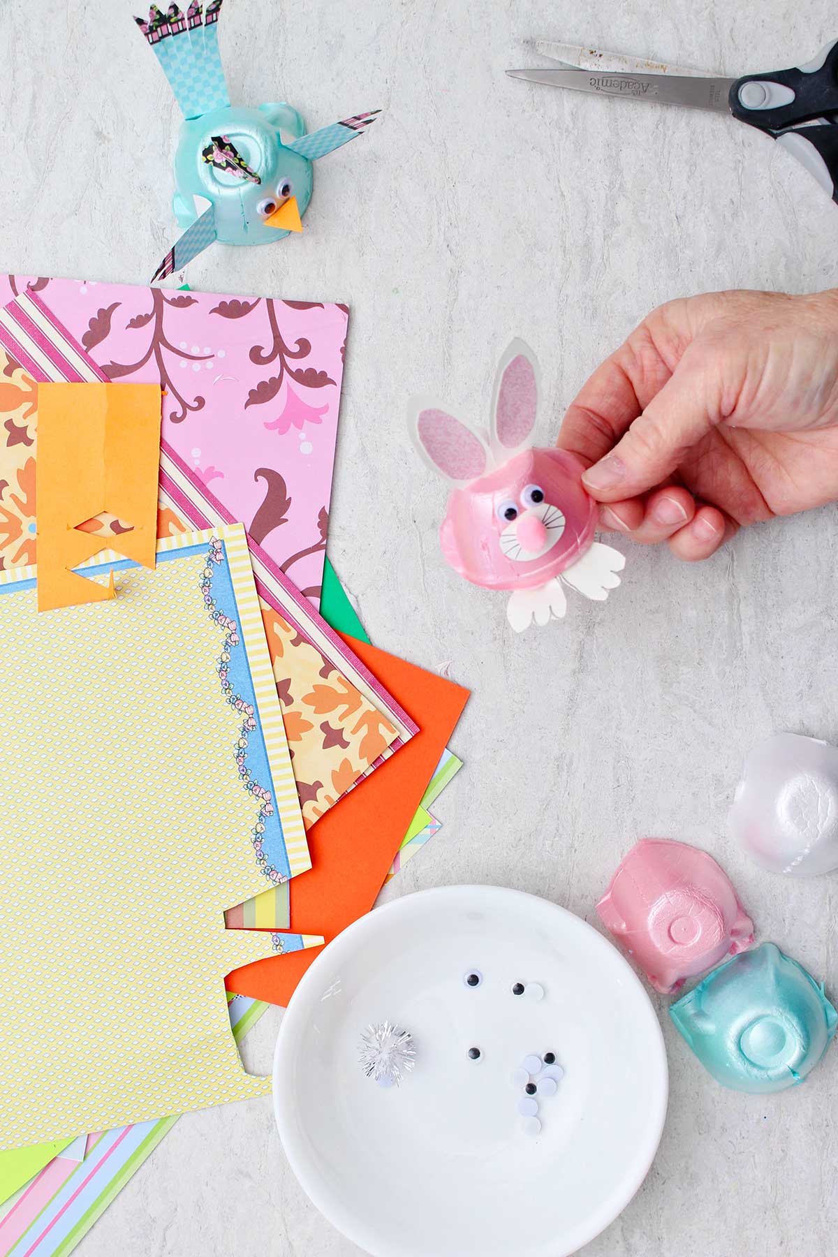 Hand holding pink bunny egg carton animal with other scraps of colorful paper and egg carton animals near by.