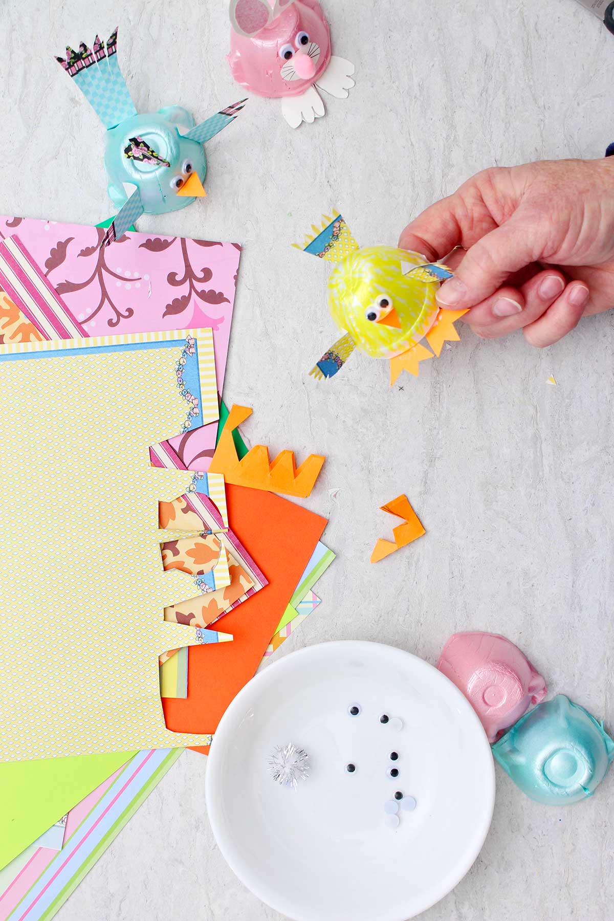 Hand holding yellow chick egg carton animal with other scraps of colorful paper and egg carton animals near by.