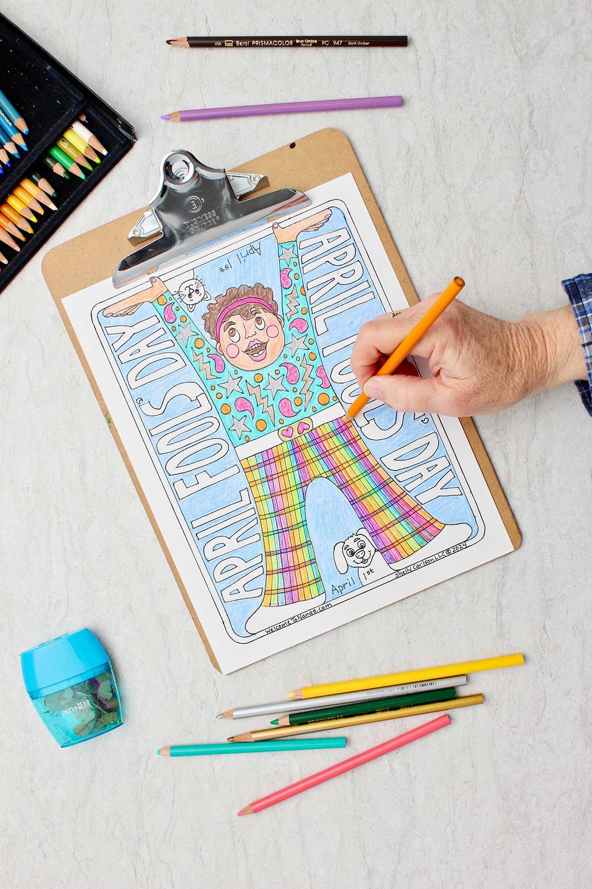 Hand coloring details in pants orange in April Fools Coloring Page clipped in clip board with colored pencils around.