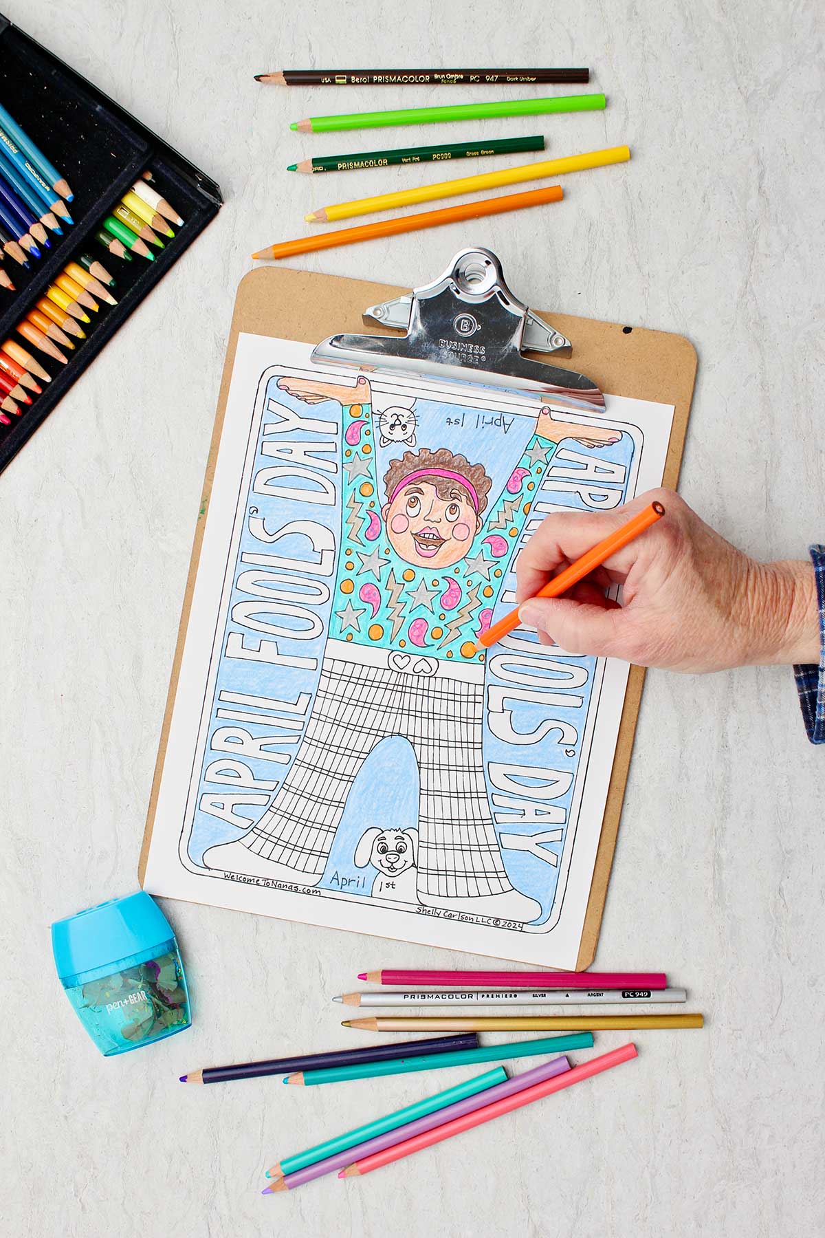 Hand coloring details in shirt orange in April Fools Coloring Page clipped in clip board with colored pencils around.