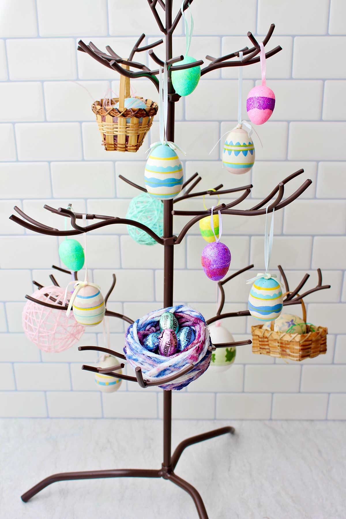 A metal tree stand with Easter decorations hanging from the branches and a colorful bird nest made of string holding candies.