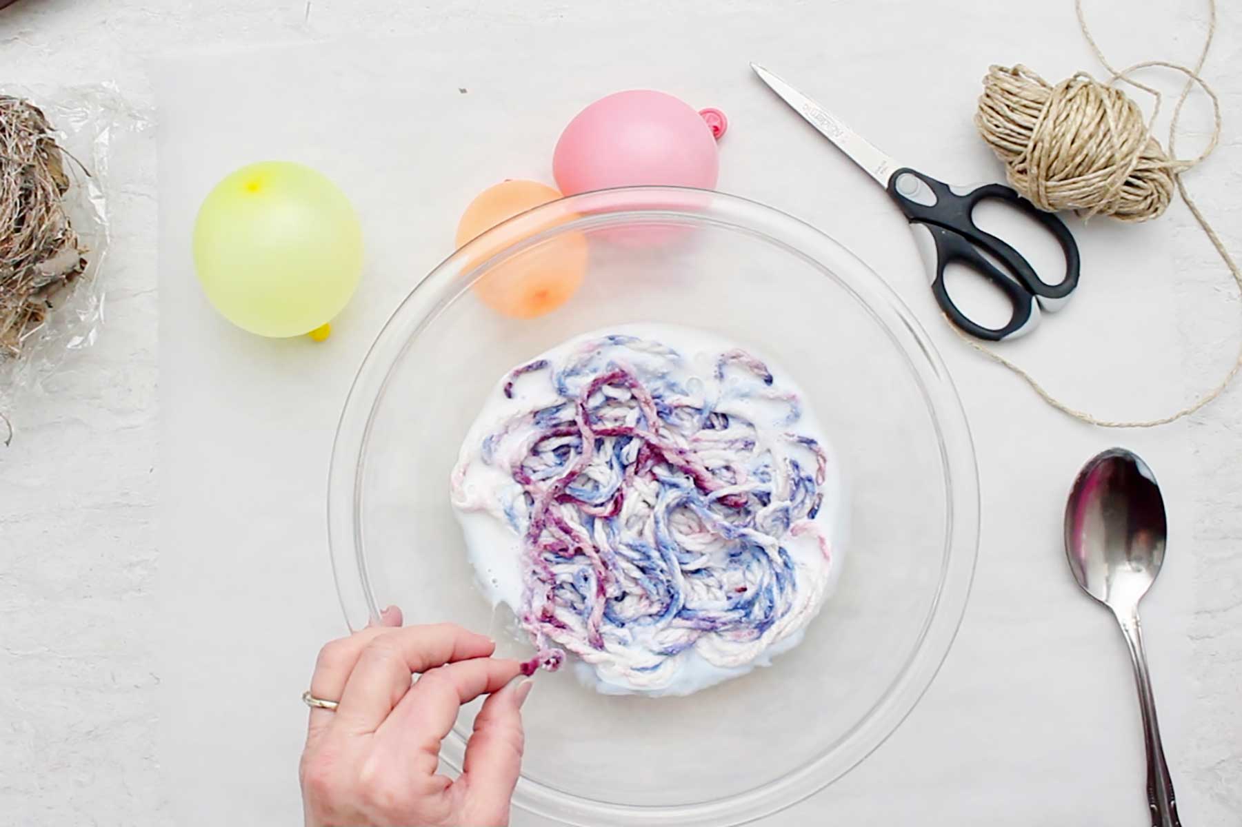 A bowl of colorful variegated yarn soaking in white glue with other supplies near by.