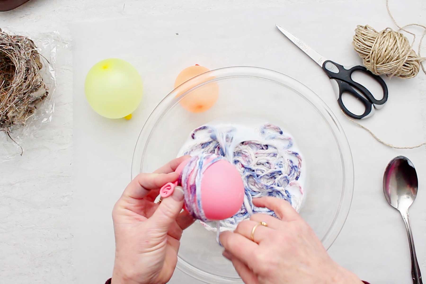 Hand winding white glue soaked yarn around a blown up pink balloon with supplies near by.